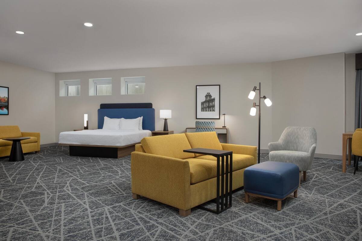 A hotel room with a bed, a sofa, a chair, a small table, and a floor lamp.