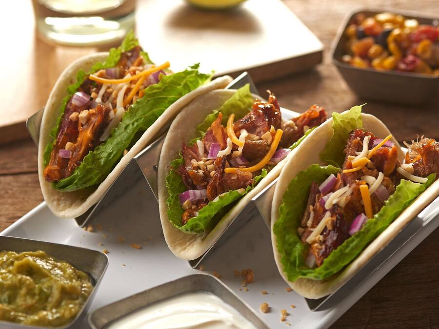 Trois tacos garnis de laitue, d’oignons rouges et de fromage râpé, présentés sur un plateau blanc, accompagnés d’un verre de boisson fraîche et de quartiers de citron vert.