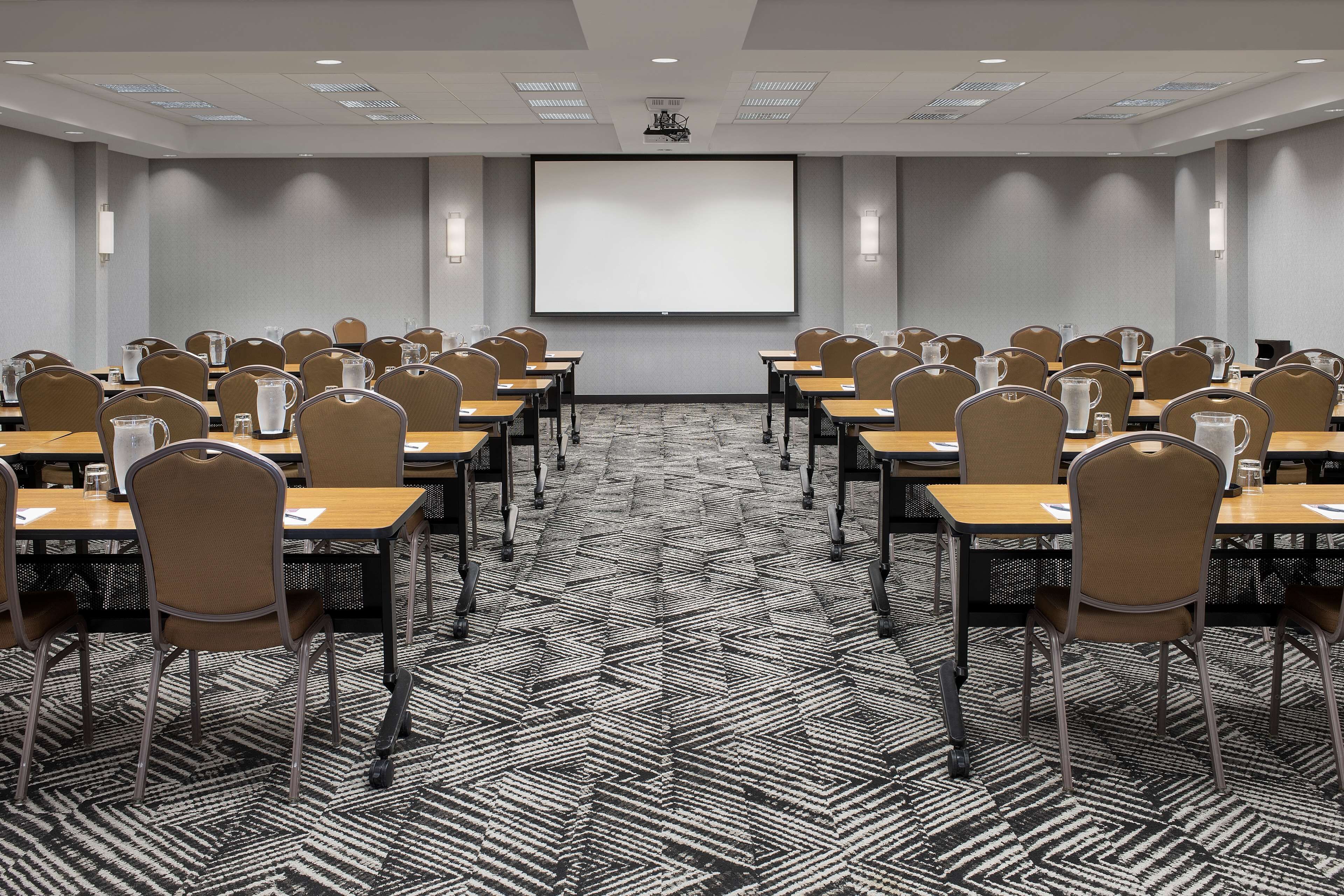 A conference room with rows of tables and chairs, a projector screen, and a patterned carpet.