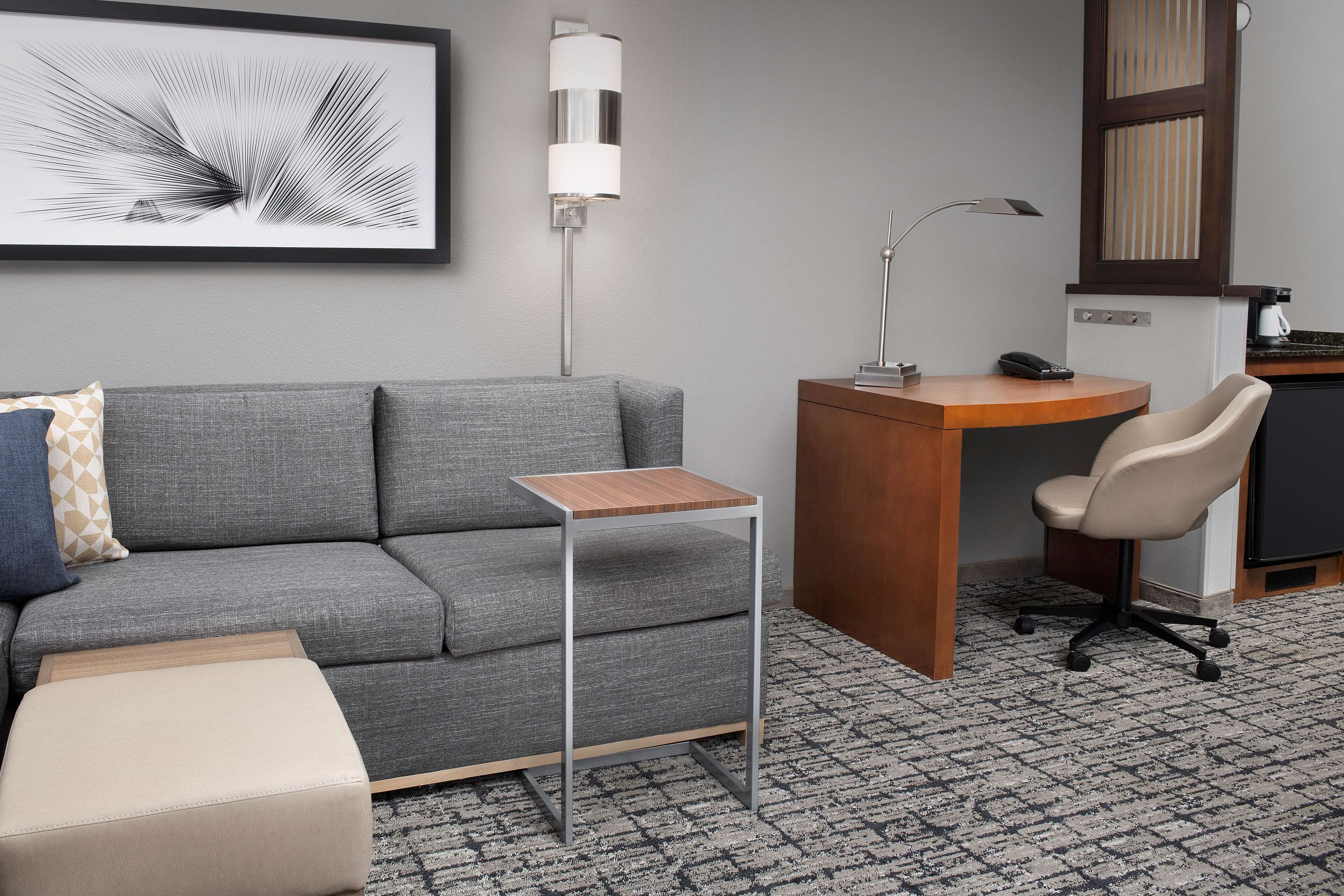 Une chambre d’hôtel moderne avec un canapé gris, une petite table d’appoint en bois, un bureau avec une chaise, un lampadaire et une œuvre d’art abstraite encadrée accrochée au mur.
