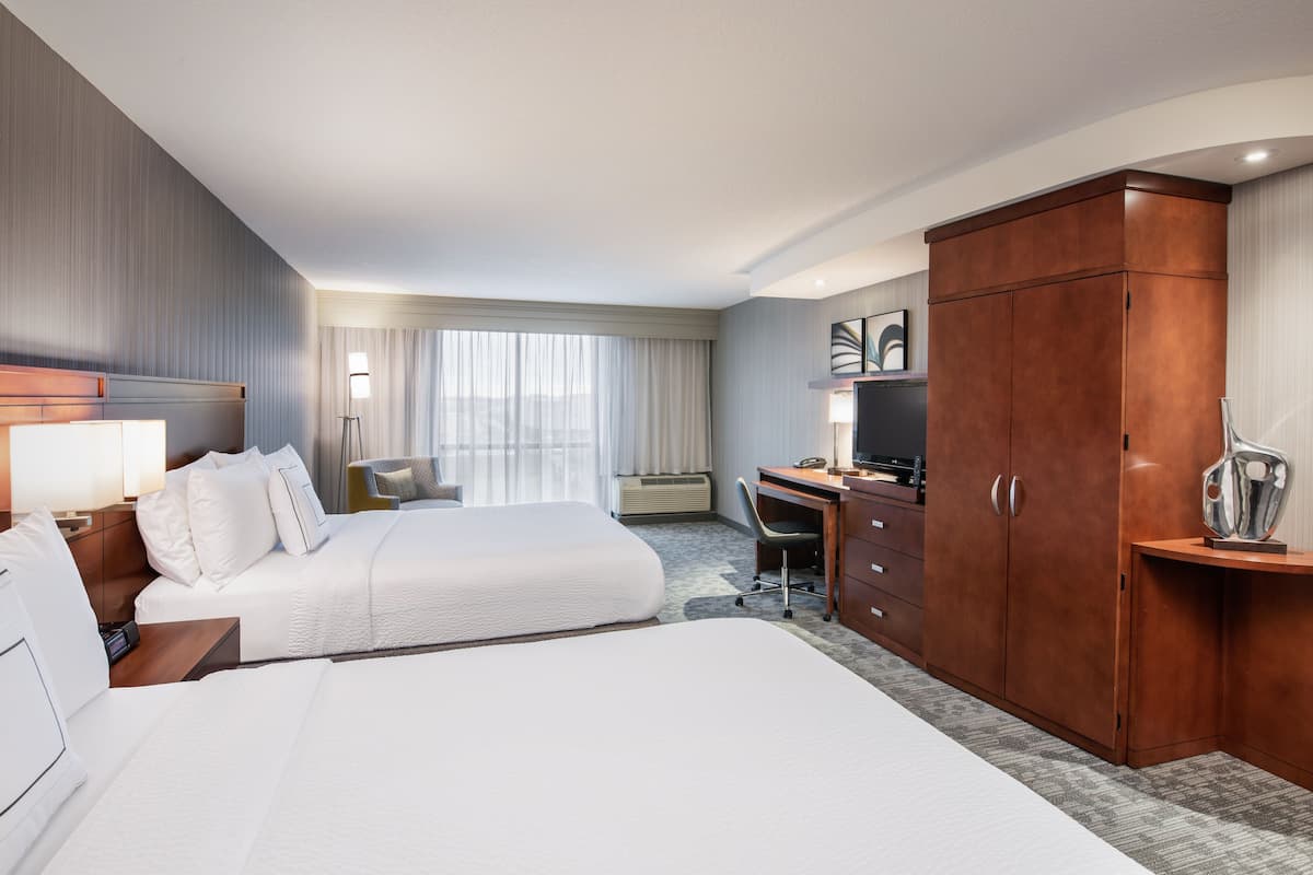 Habitación de hotel con dos camas, un escritorio, un armario, un televisor y un ventanal grande con cortinas.