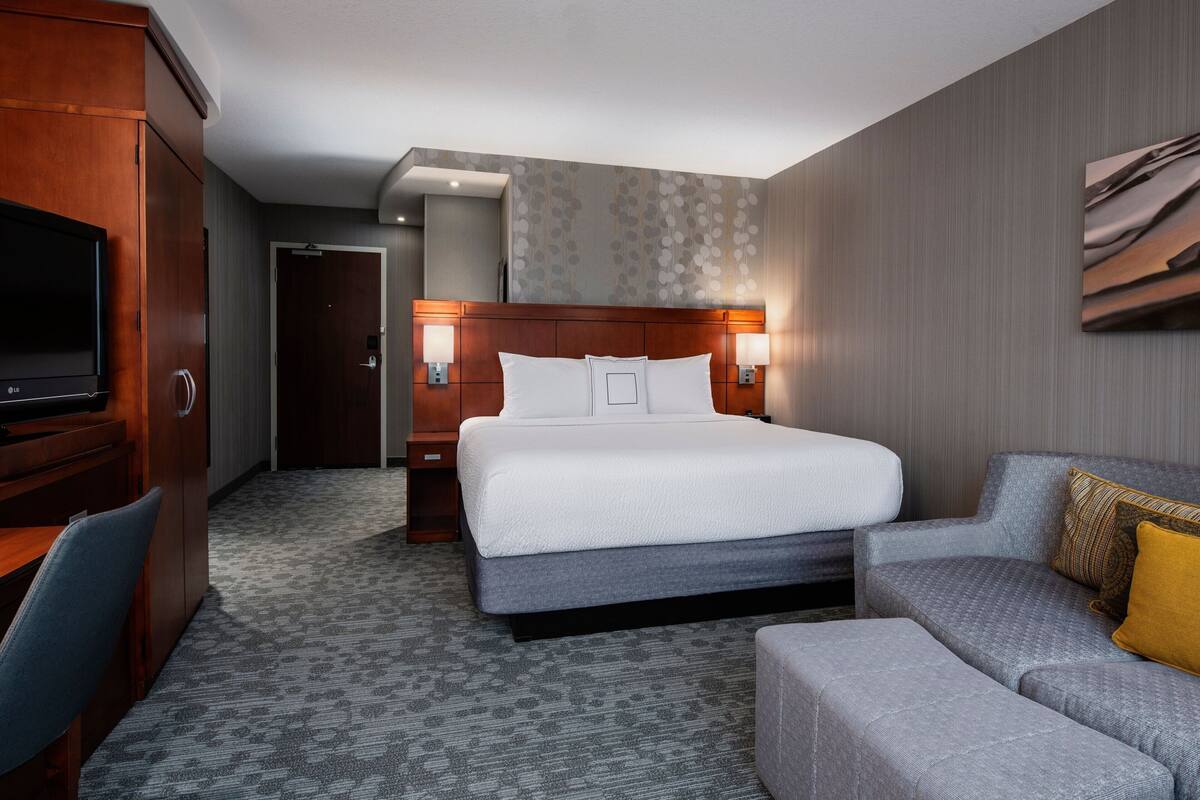 Quarto de hotel com uma cama grande, um sofá cinzento, uma televisão e um guarda-roupa de madeira.