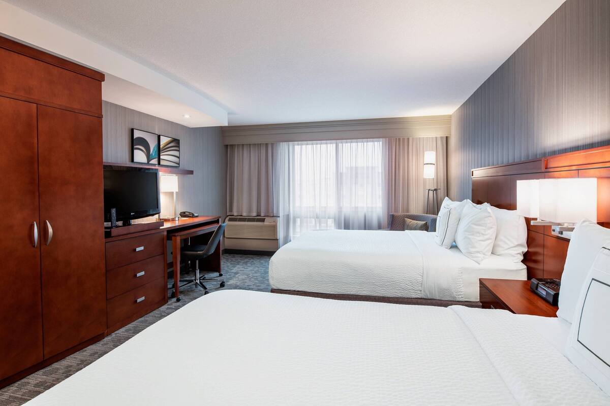 Quarto de hotel com duas camas, uma escrivaninha, televisão e uma janela grande com cortinas.