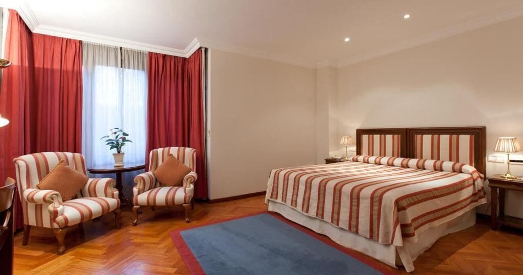 Habitación de hotel con una cama grande, dos sillones, una mesita con una planta y una ventana con cortinas rojas.