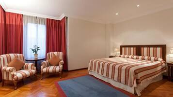 Habitación de hotel con una cama grande, dos sillones, una mesita con una planta y una ventana con cortinas rojas.