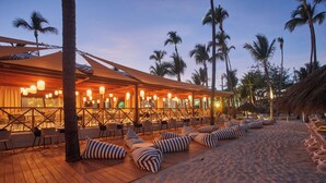 A beachside resort with wooden deck seating, palm trees, and a covered dining area.