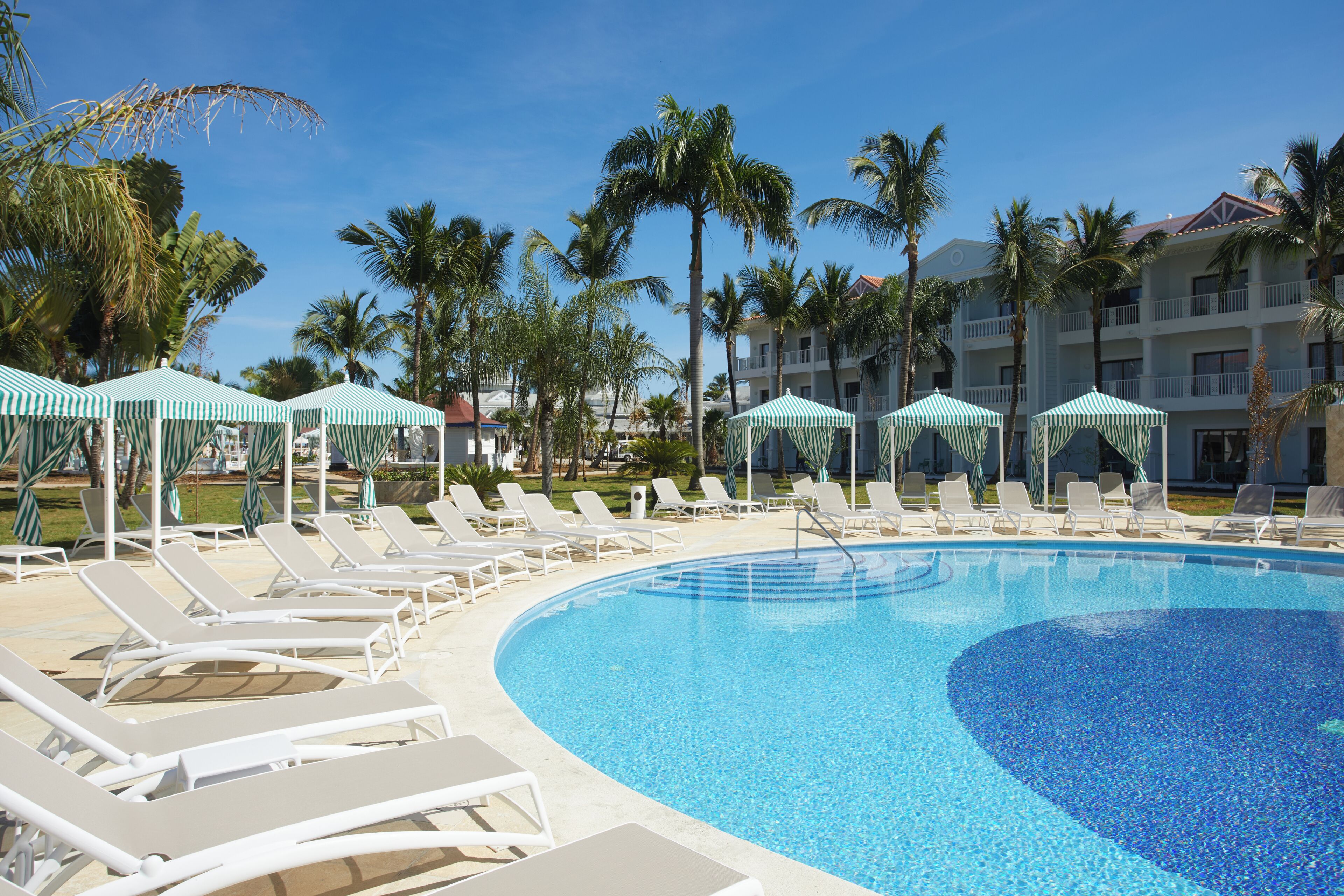 Une piscine entourée de palmiers et d’un bâtiment d’hôtel, avec des chaises longues et des parasols.