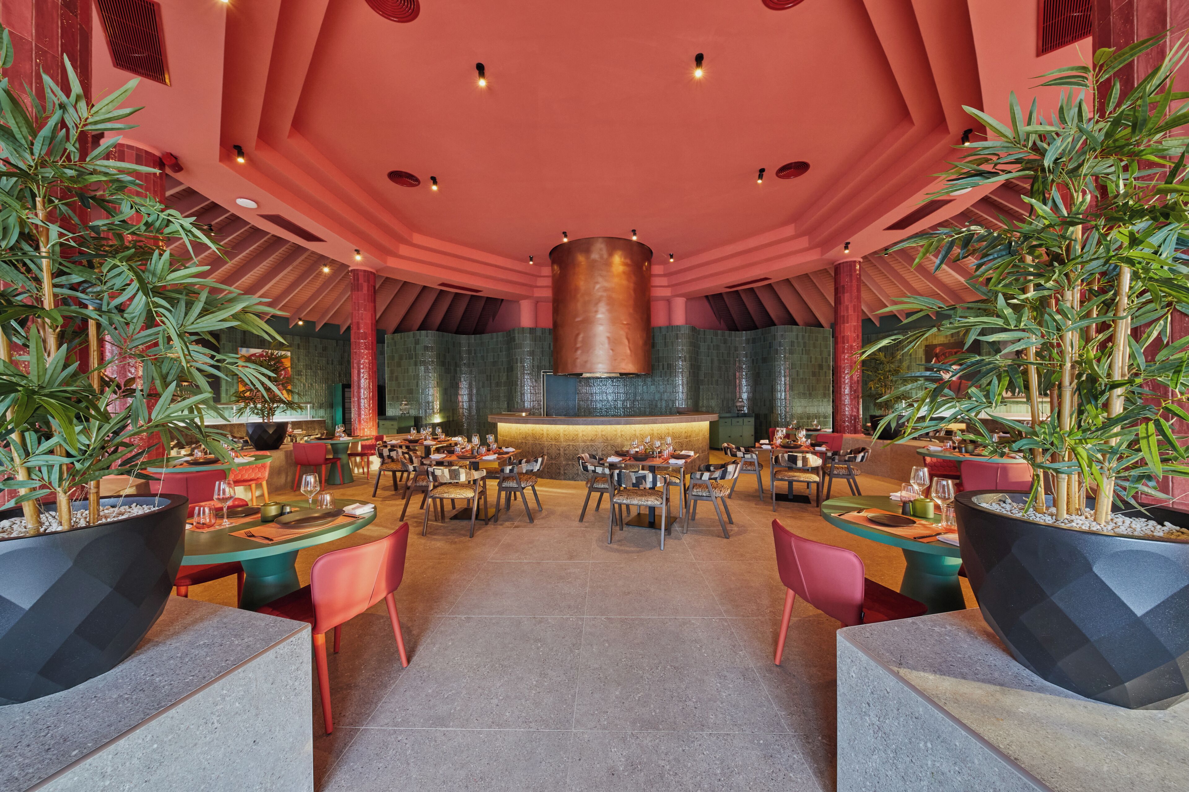 Un restaurant doté de nombreuses tables et chaises, d’un pilier décoratif central et de plantes en pot.