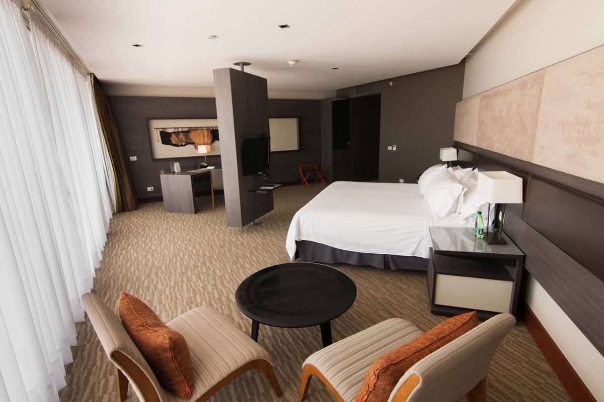 Una habitación de hotel con una cama grande, un escritorio, una silla, una mesita y una mesita de noche con una lámpara.