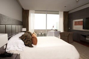 A hotel room with a large bed, a television, a desk with a lamp, and a view of the city through large windows. - Enjoy Antofagasta (Antofagasta)