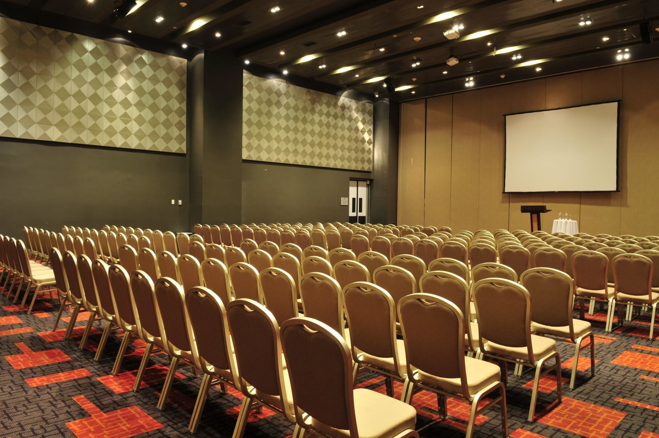 A conference room with rows of chairs, a projector screen, and a patterned wall.