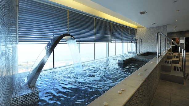 Una piscina interior moderna con caída de agua, amplias ventanas y una escalera que lleva a un nivel superior.