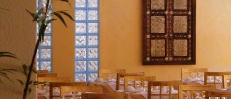 A dining area with tables set for service, a large window with blue panes, and a decorative wooden panel on the wall.