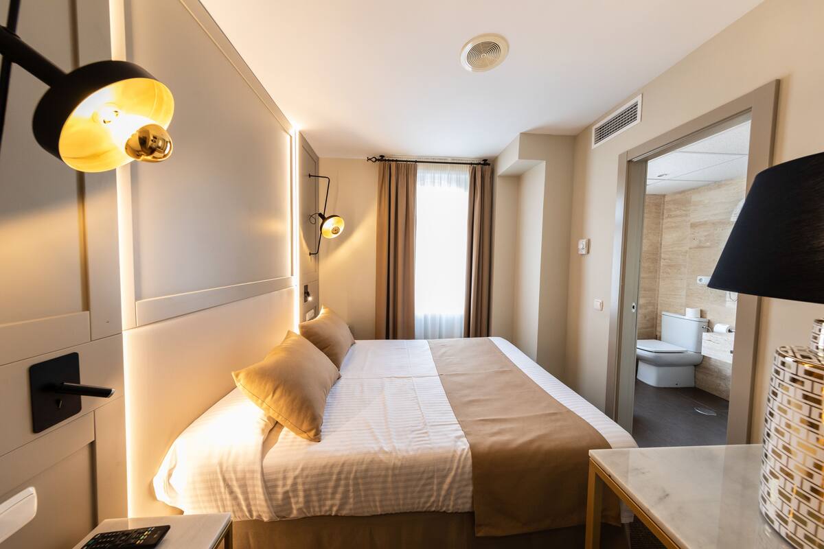 Una habitación de hotel con cama, mesita de noche, lámpara y vistas al baño.