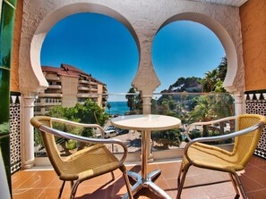 A balcony with two wicker chairs and a small table, overlooking a coastal view with buildings and greenery.