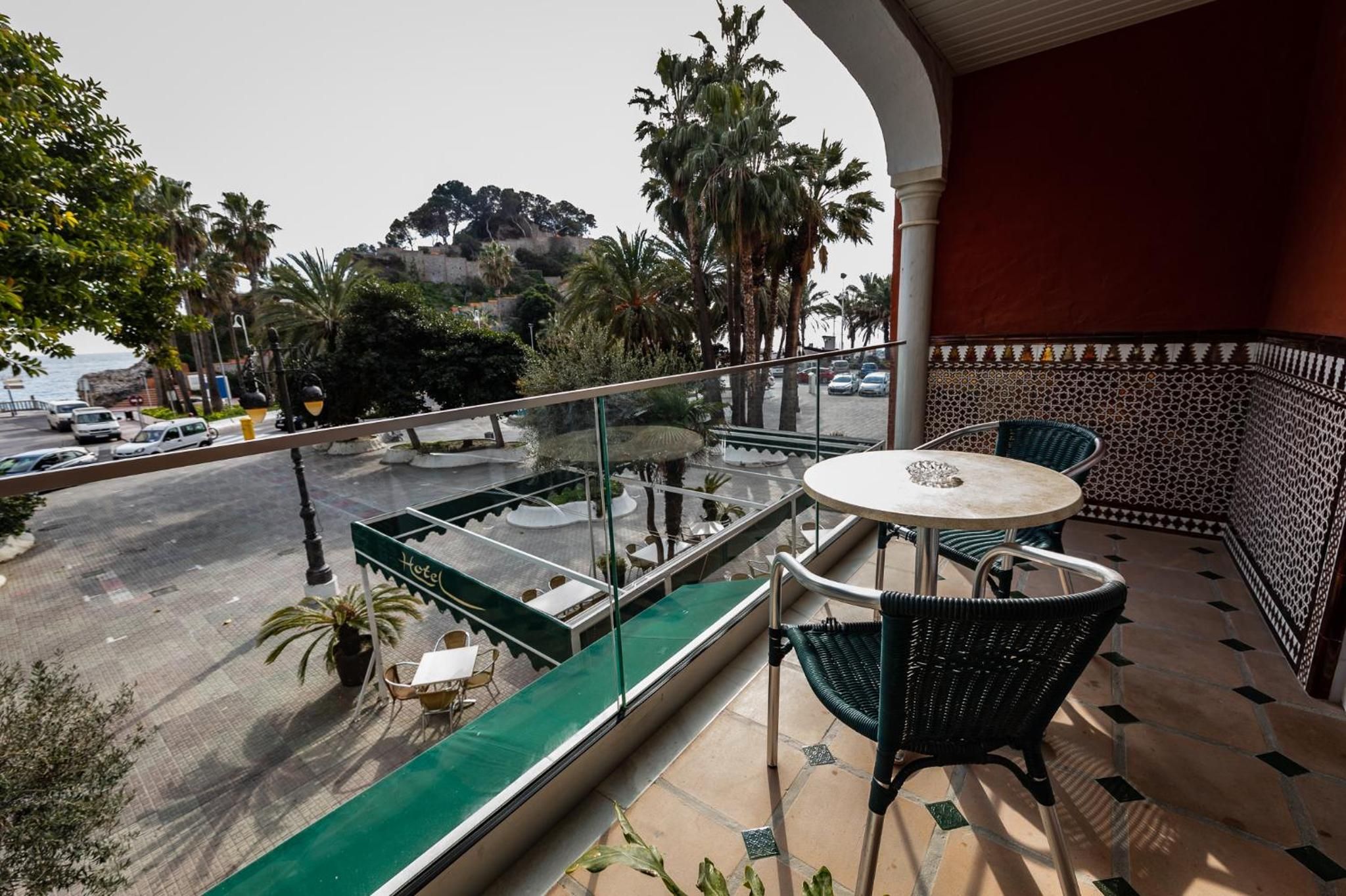 A balcony with a glass enclosure, a round table, and chairs, overlooking a street with cars and palm trees.