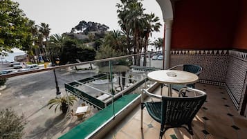 Un balcon doté d’une paroi vitrée, une table ronde et des chaises, donnant sur une rue animée où l’on peut voir des voitures et des palmiers.