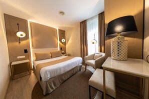 A hotel room with a large bed, bedside tables, a desk with a lamp, and a chair.