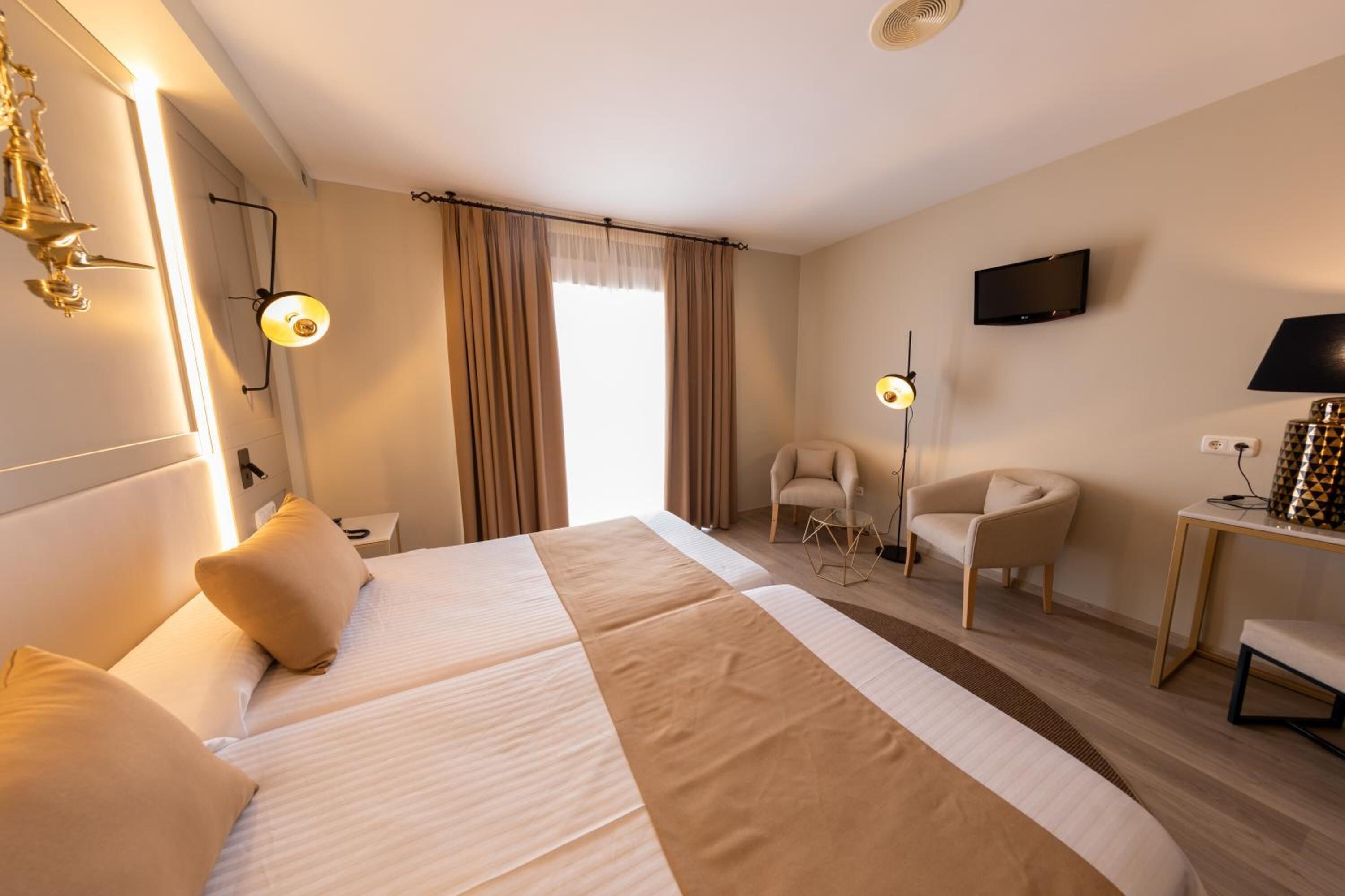 A hotel room with a large bed, two bedside tables, a desk, a chair, a TV, and a window with curtains.