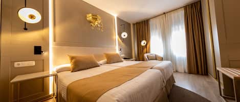 A hotel room with two beds, a bedside table, a lamp, a mirror, and a window with curtains.