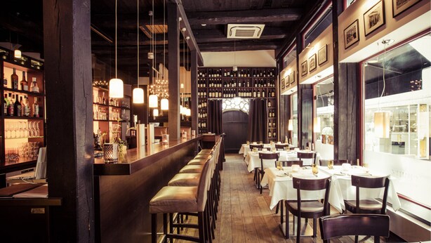 A bar area with a long counter, multiple stools, and a wine rack filled with bottles. Tables with chairs are arranged in the dining area. - Altera Hotel im Herbartgang (Oldenburg)