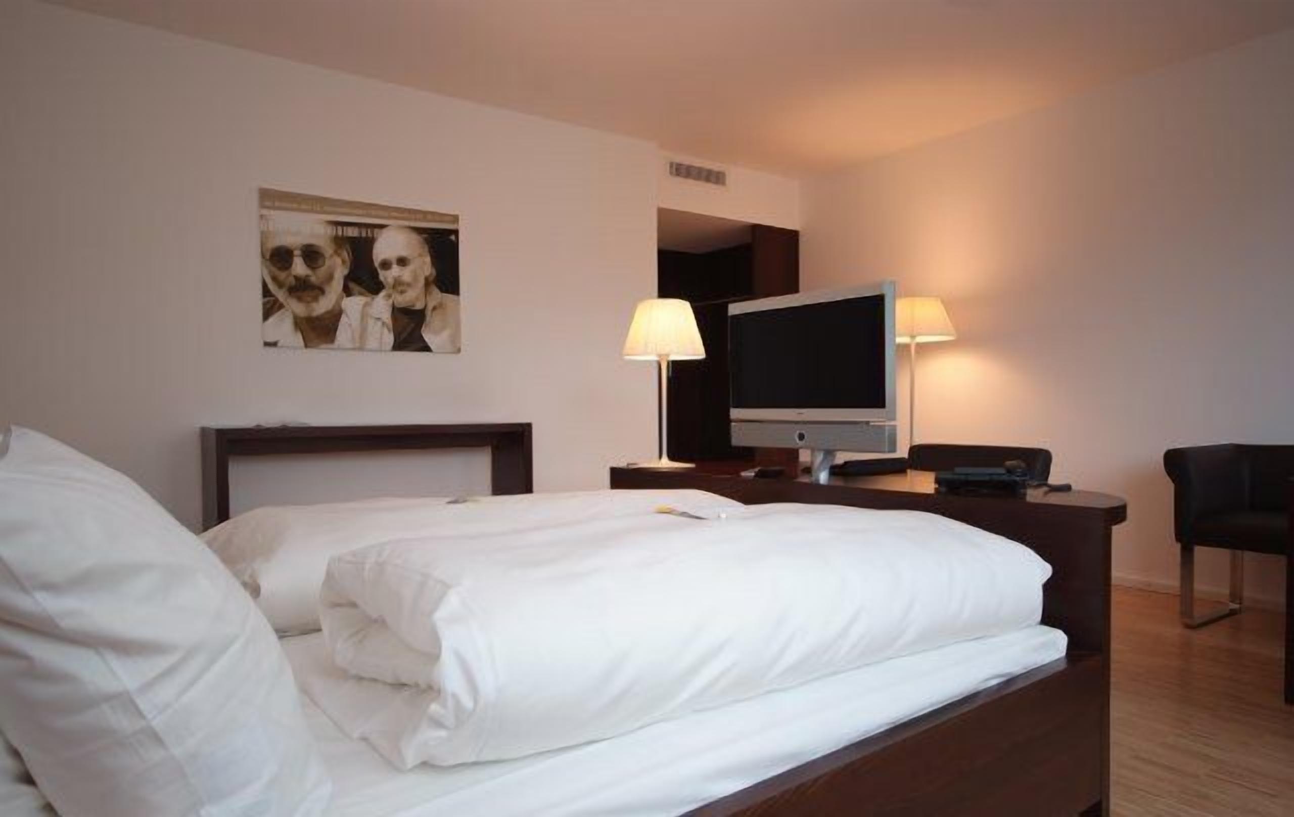 Una habitación de hotel con una cama, televisión, escritorio con lámpara y una silla.