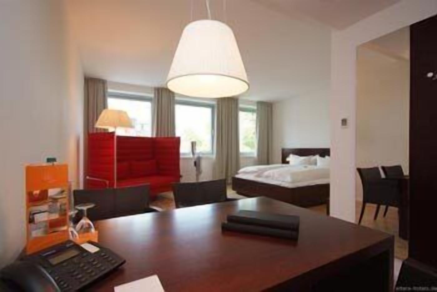 A hotel room with a bed, a desk with a telephone, a red sofa, and a lamp.