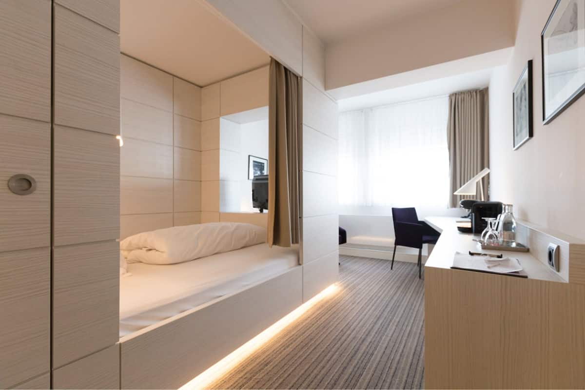 A modern hotel room with a bed, a desk, a chair, and a large window with curtains.