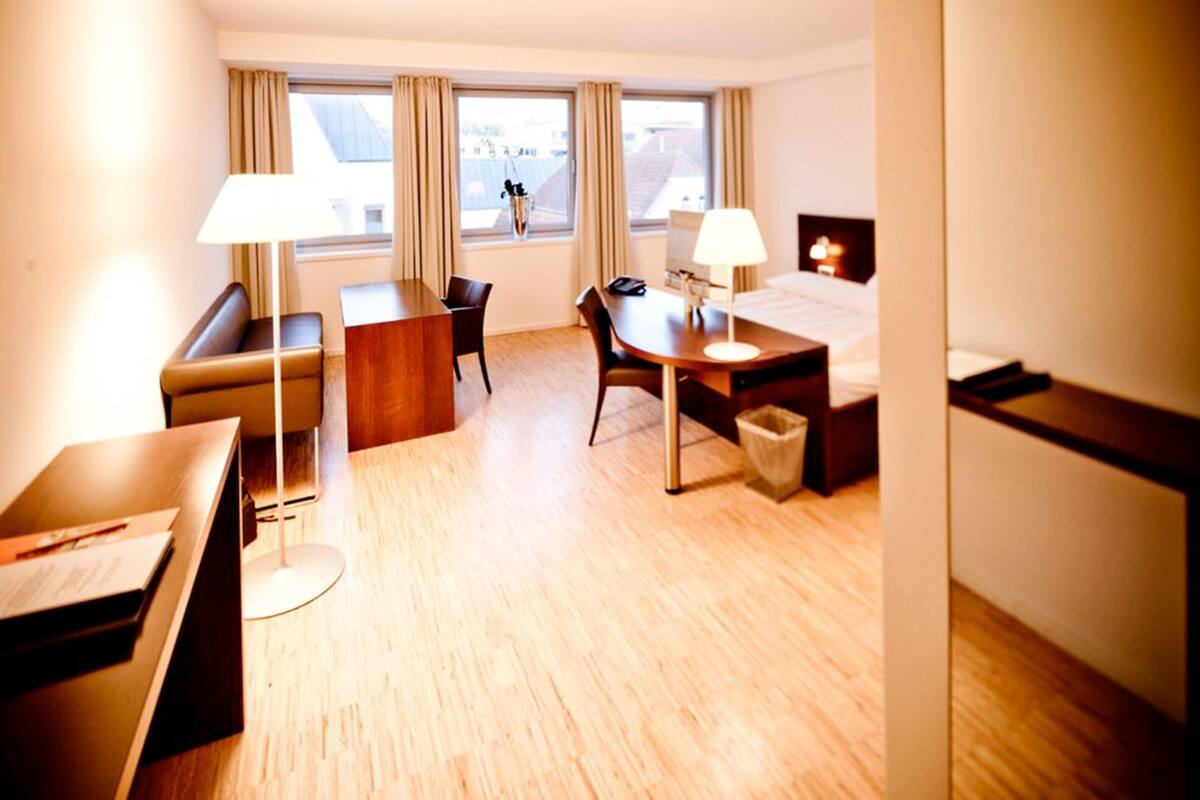 A hotel room with a bed, a desk, a chair, a sofa, a lamp, and a trash bin.