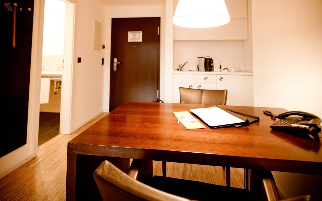 Habitación de hotel con mesa de comedor de madera, una silla, un teléfono y una pequeña zona de cocina con microondas y cafetera.