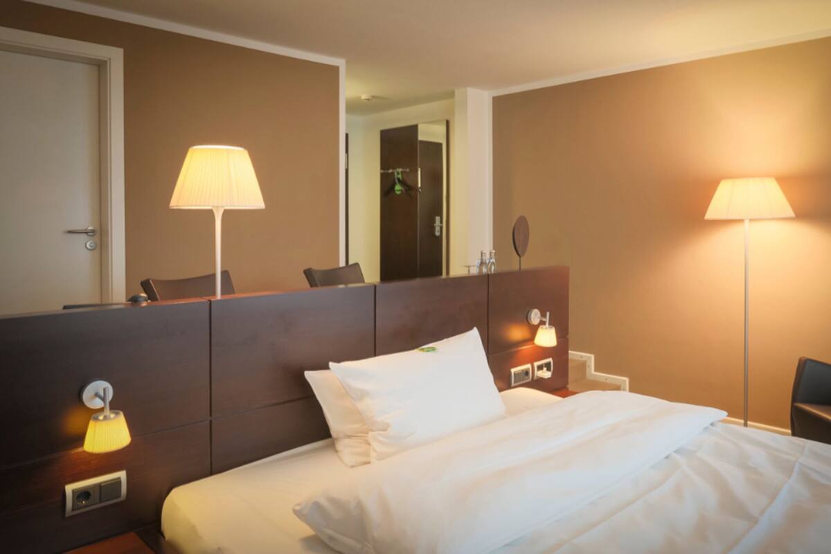 A hotel room with a bed, two bedside lamps, a mirror, a chair, and a door.