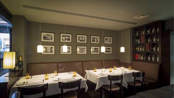A restaurant interior with multiple tables set for dining, framed pictures on the wall, and a bookshelf with various items.