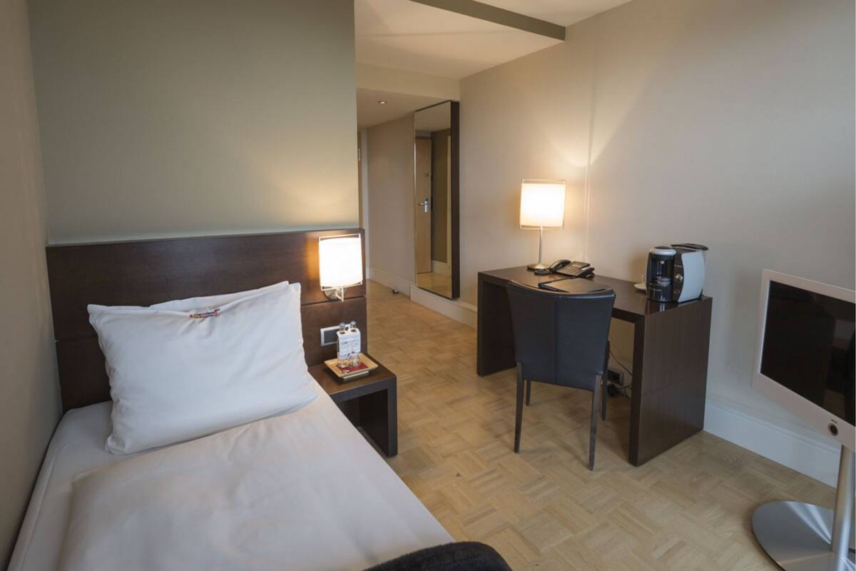 A hotel room with a bed, bedside table, desk, chair, and a TV.