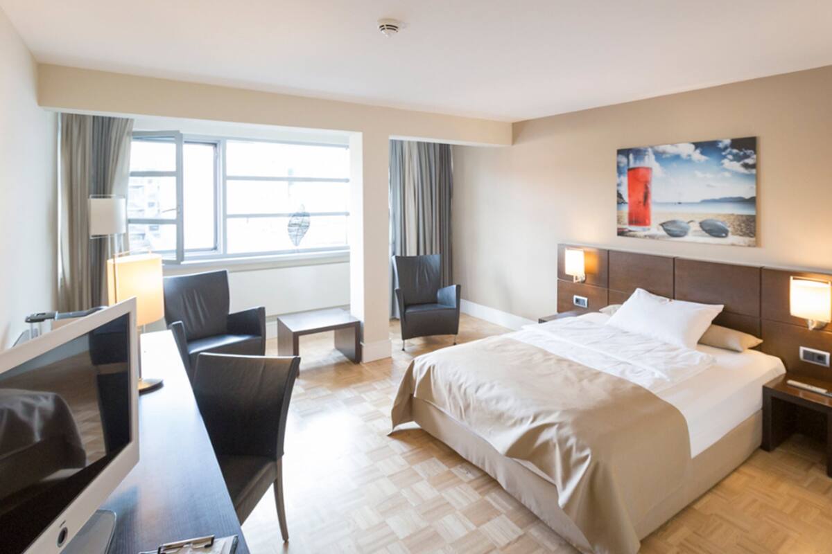 A hotel room with a large bed, a desk with a computer, two chairs, a TV, and a painting on the wall.