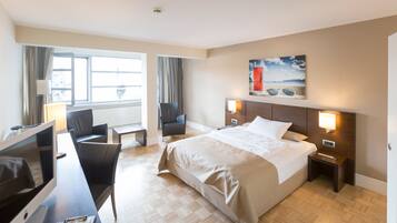 A hotel room with a large bed, a desk with a computer, two chairs, a TV, and a painting on the wall.