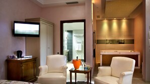 A hotel room with a bed, two armchairs, a television, a small table with a bottle and glasses, and a view into the bathroom and kitchen. - Andris Hotel (Ercolano)