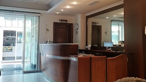 A hotel lobby with a reception desk, a large mirror, a clock, and a view of the outside through a glass door. - Andris Hotel (Ercolano)