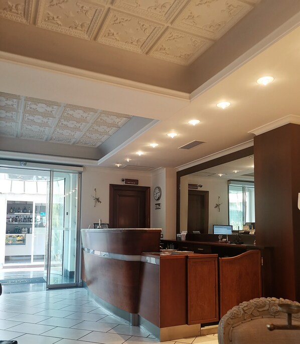 A hotel lobby with a reception desk, a large mirror, a clock, and a view of the outside through a glass door. - Andris Hotel (Ercolano)