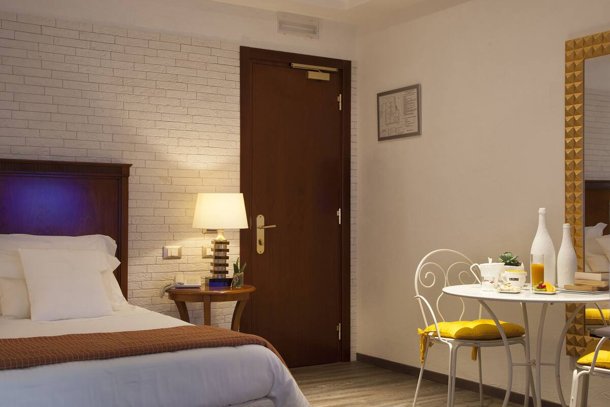 Hotellihuone, jossa on sänky, yöpöytä lampun kanssa, pieni pöytä aamiaistarvikkeita varten ja ovi.