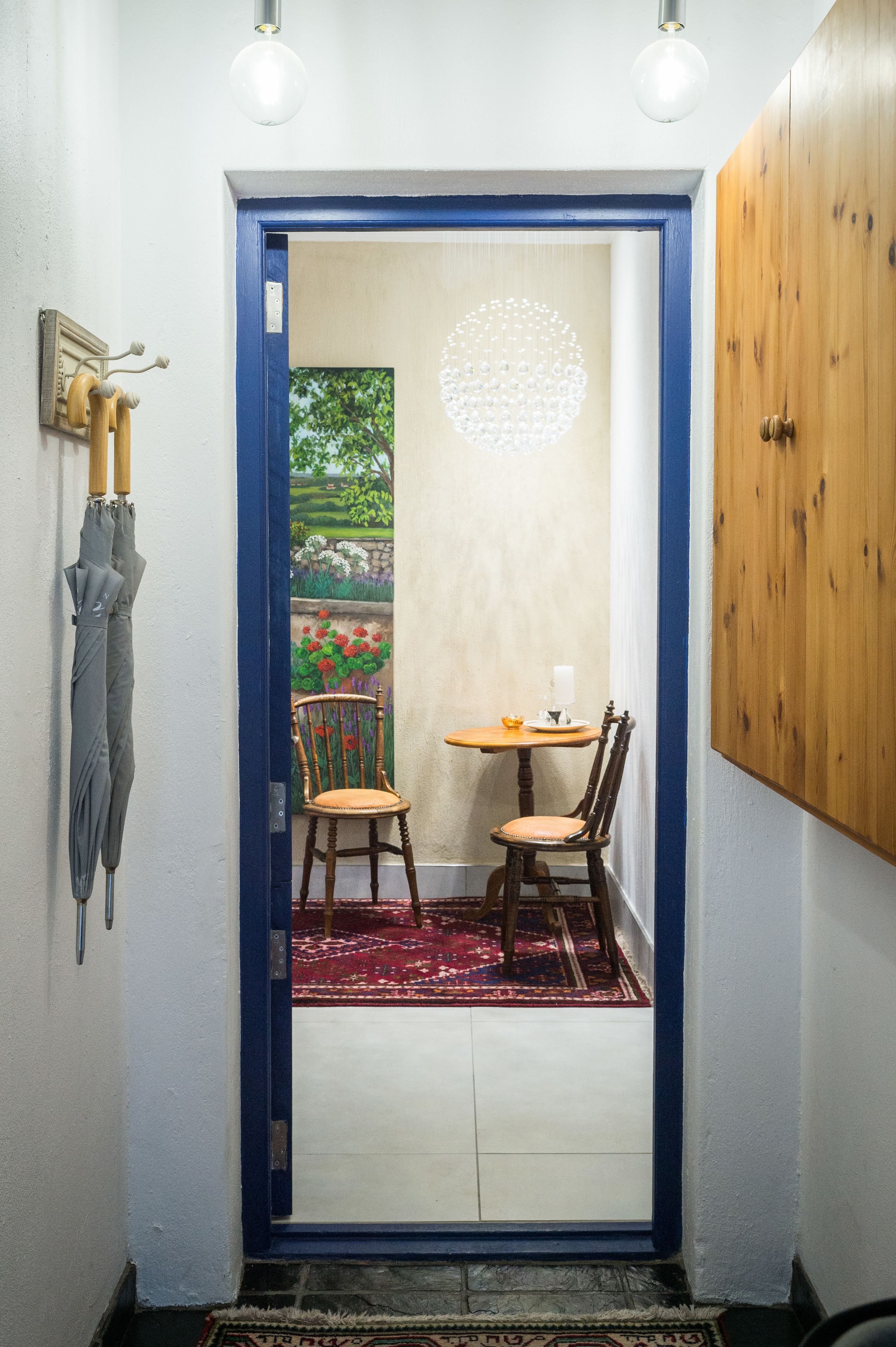 A narrow hallway with a blue-framed door leading to a room with a small table and two chairs, a rug, and a wall with a circular light fixture.