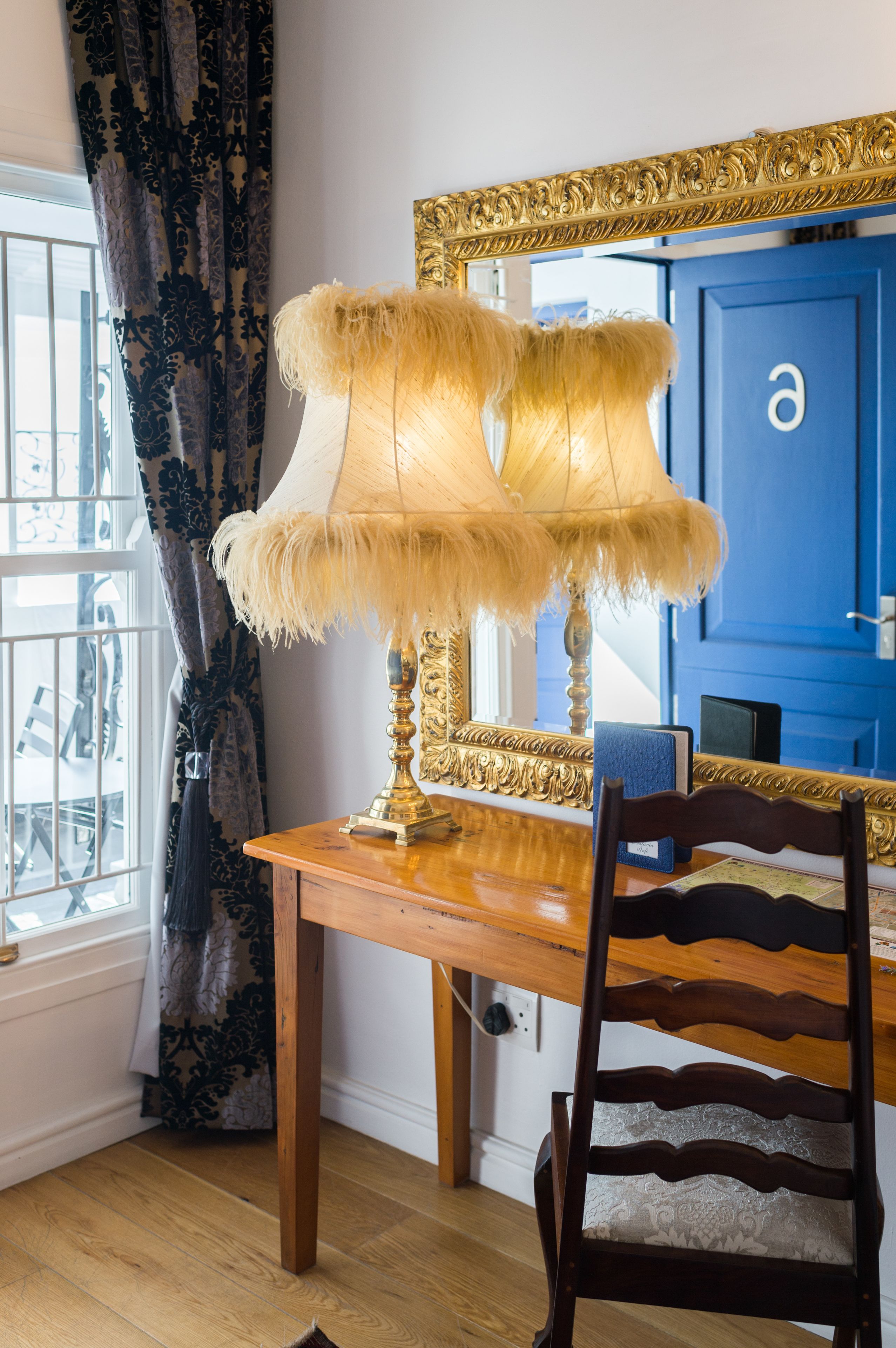 A room with a wooden table, a chair, a lamp with a feathered shade, a blue door, and a window with curtains.