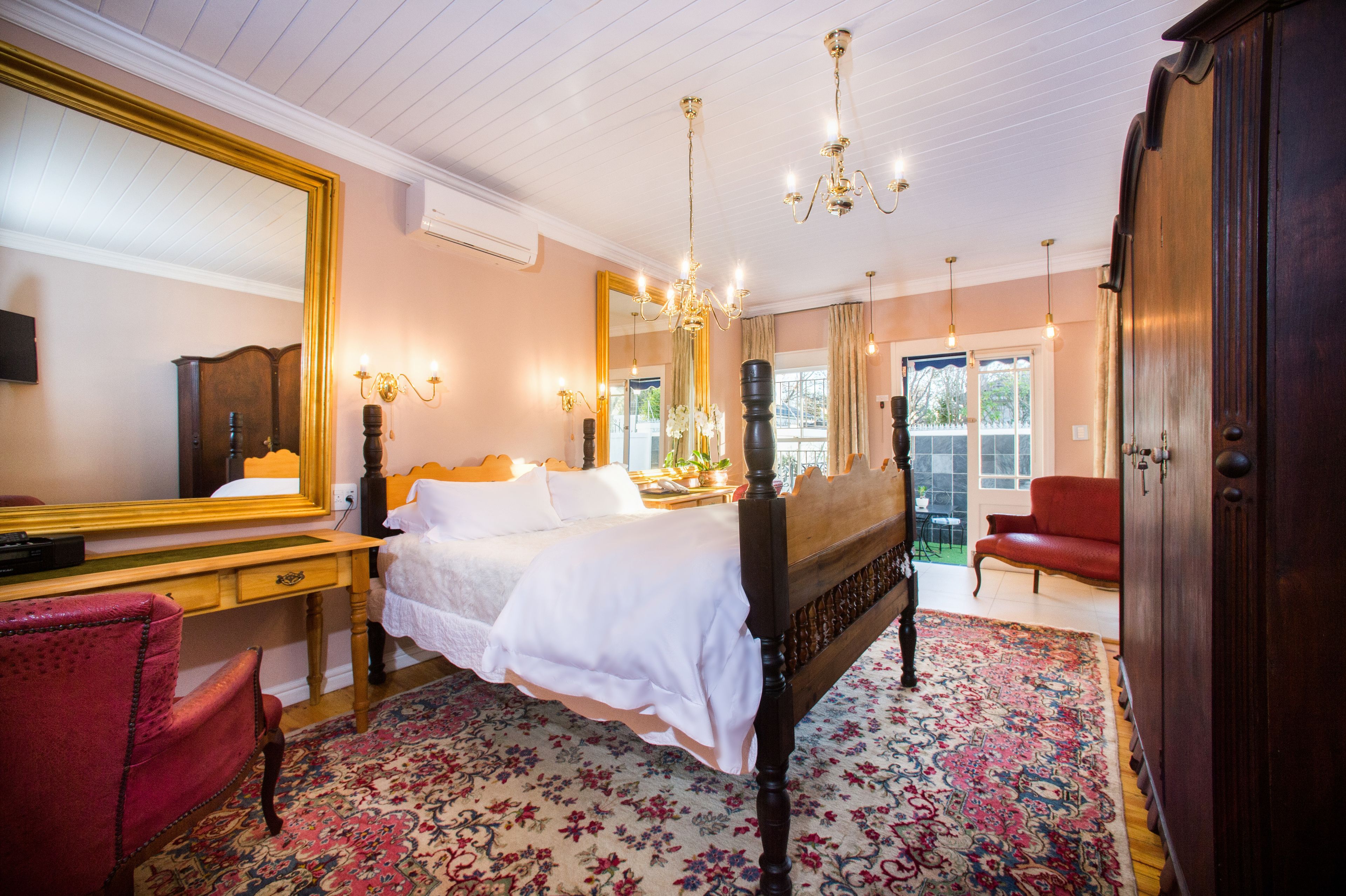 A bedroom with a large bed, a wooden desk, a red chair, a large mirror, and a chandelier.