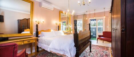 A bedroom with a large bed, a wooden desk, a red chair, a large mirror, and a chandelier.