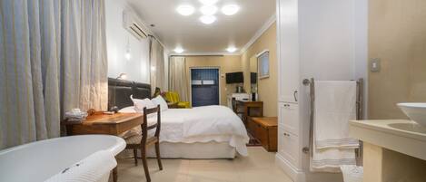 A hotel room with a bathtub, a bed, a desk, a chair, a television, and a bathroom with a sink and mirror.