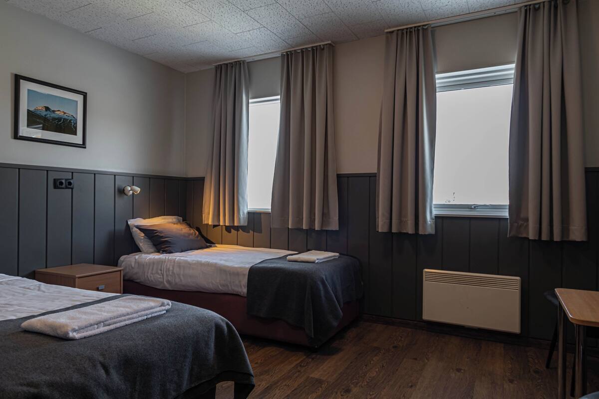 A hotel room with two beds, a bedside table, a chair, a radiator, and a window with curtains.