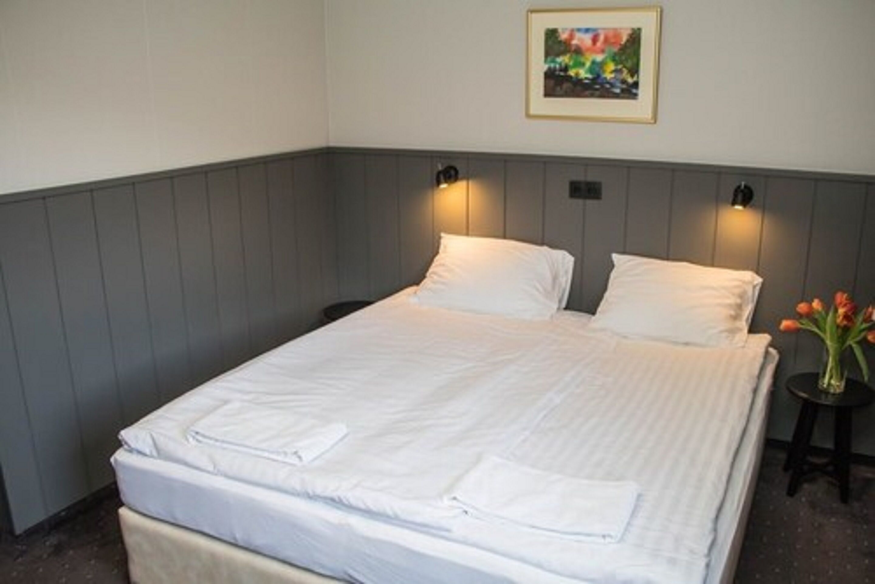 Una cama bien hecha con sábanas y almohadas blancas, una mesita de noche con un jarrón de flores, y un cuadro en la pared.