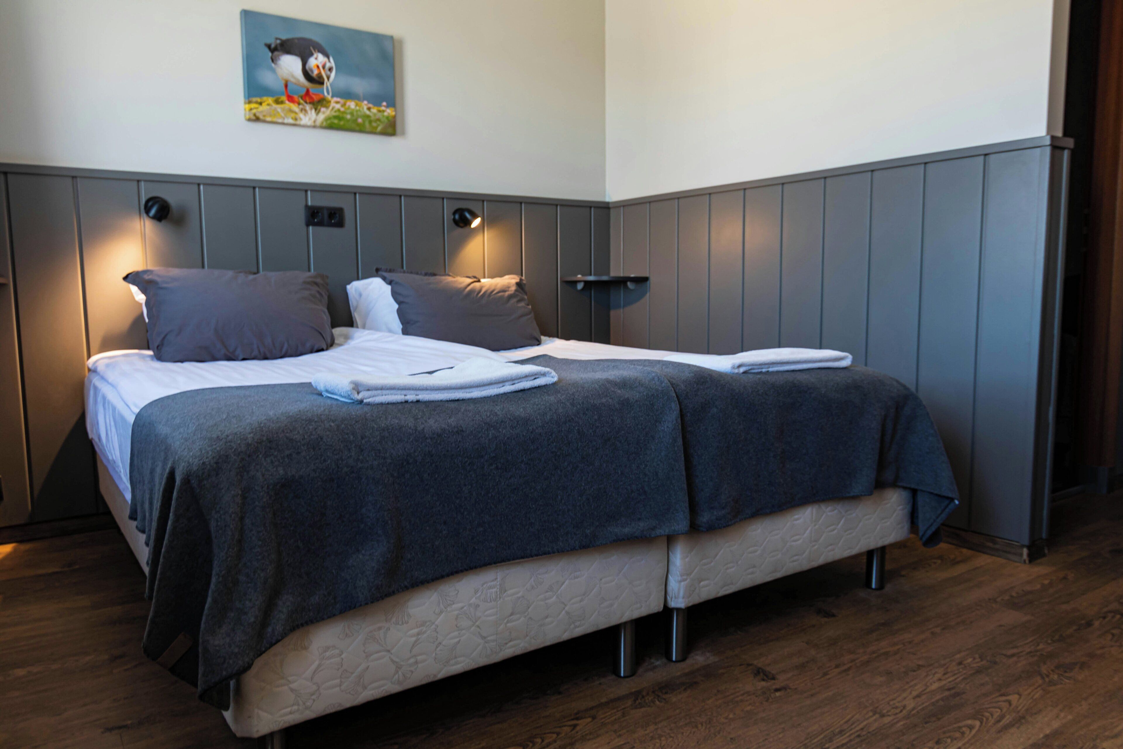 A double bed with a grey duvet and pillows, a bedside table with a lamp, and a wall-mounted picture of a bird.