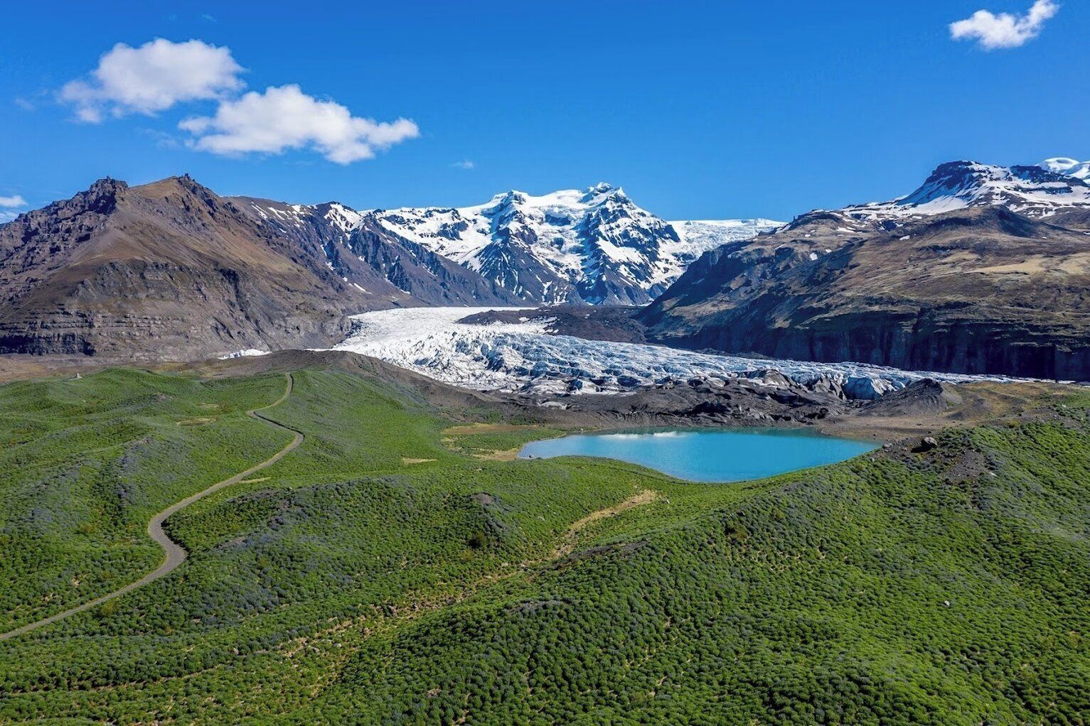 A glacier with a small lake in the foreground, surrounded by mountains and greenery.