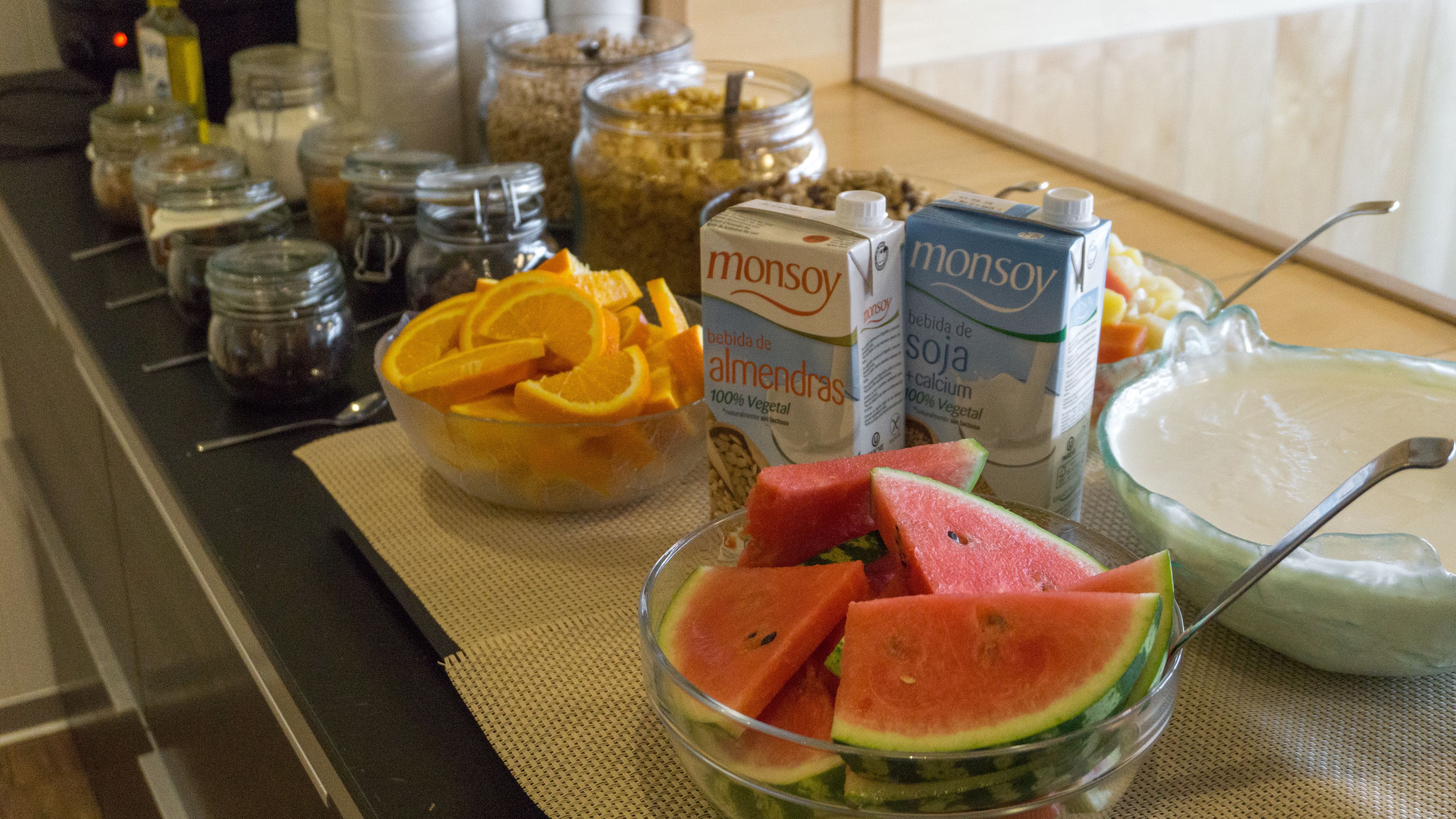 A kitchen counter with various food items and containers, including a bowl of sliced oranges, a bowl of watermelon, and cartons of milk.