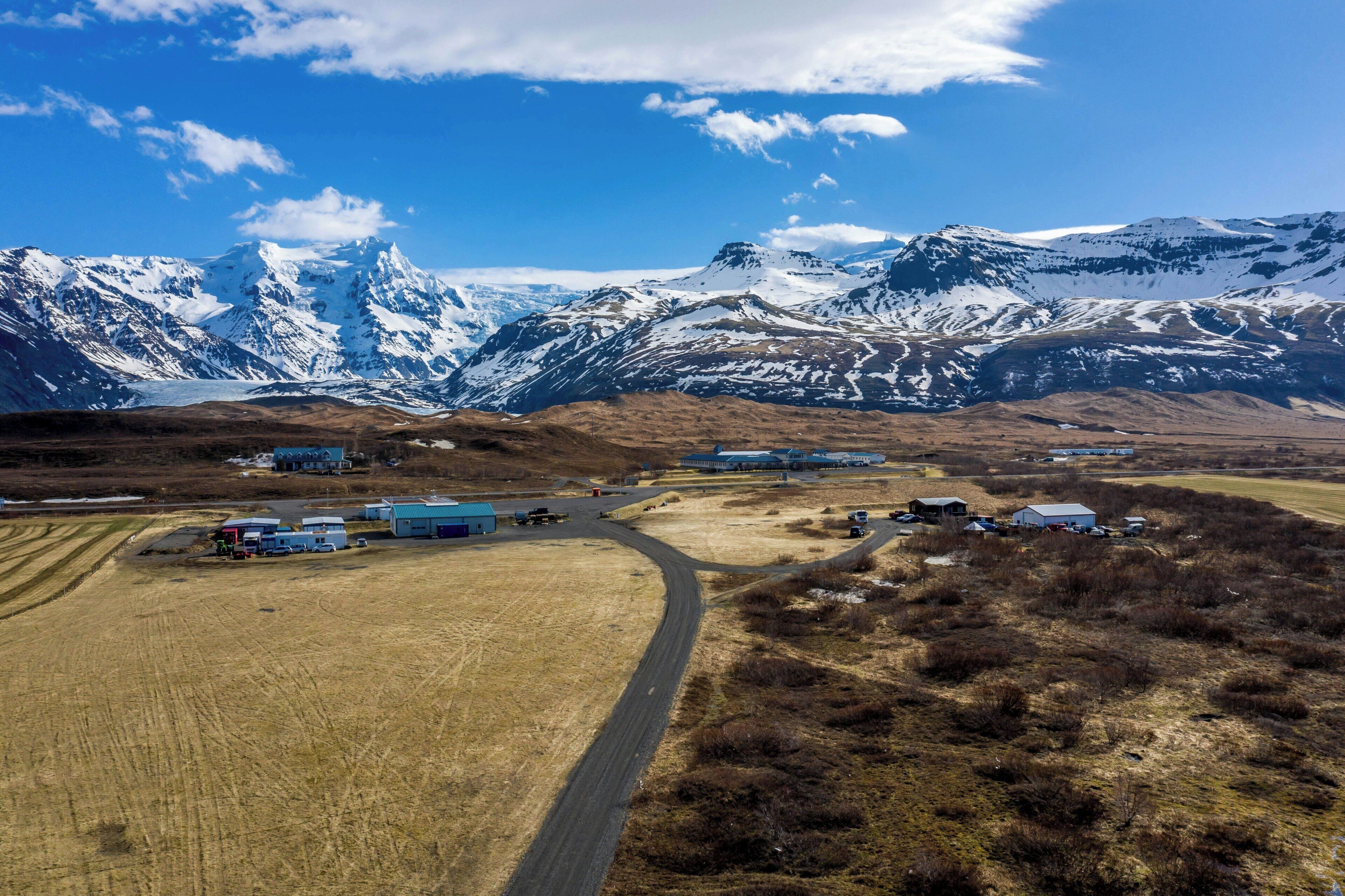 A rural landscape with a road leading to a cluster of buildings, surrounded by snow-capped mountains.